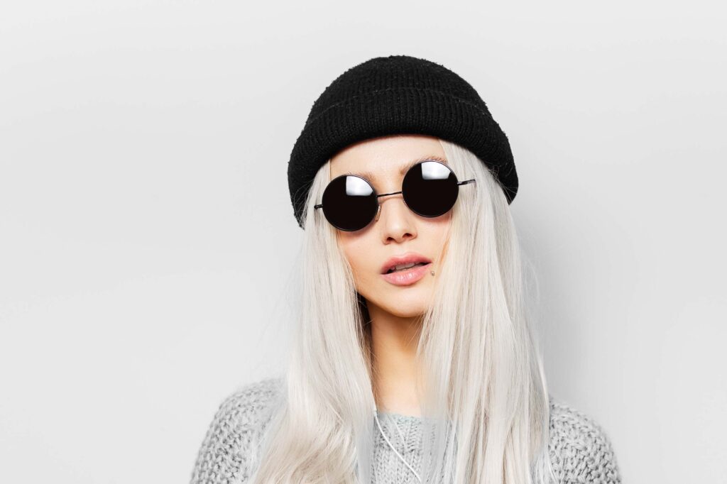 Eine Person mit langem, platinblondem Haar trägt eine schwarze Strickmütze, eine runde schwarze Sonnenbrille und einen grauen Pullover vor einem schlichten weißen Hintergrund - ein stilvoller Look, der perfekt zum Friseursalon Black Hair & Beauty für Kinder und Teenager passt.