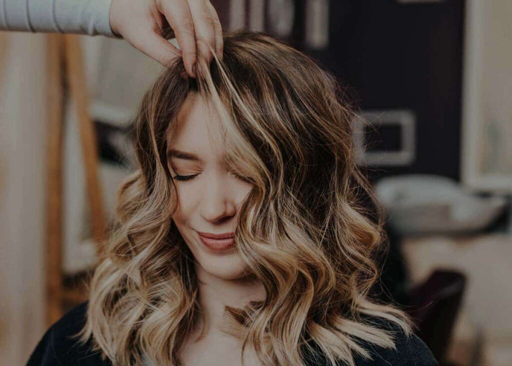 Eine Person mit gewelltem, schulterlangem Haar sitzt mit geschlossenen Augen da, während eine andere Hand die vordere Partie stylt - ein ruhiger Moment bei Coiffeur Frauen Black Hair & Beauty Arlesheim.