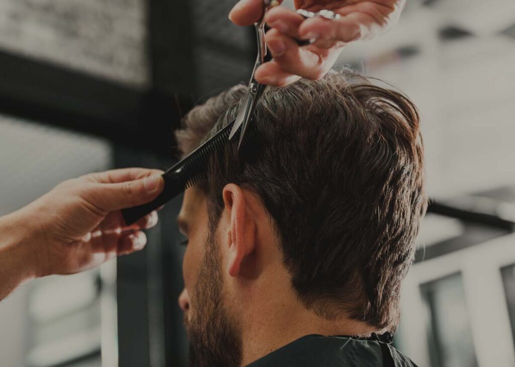 Eine Person lässt sich bei einem Coiffeur für Männer die Haare schneiden; ein Stylist benutzt Schere und Kamm, um das braune Haar am Hinterkopf zu stutzen und zeigt dabei echte Black Hair & Beauty Expertise.